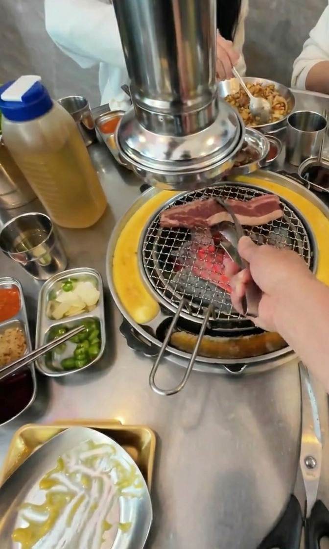 Una mano con pinze di metallo adagia strisce di carne di manzo su una griglia a carbone rovente circondata da contorni.