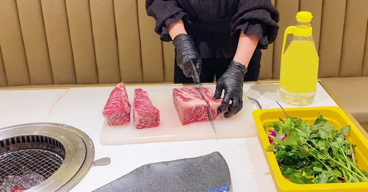 Una persona con guanti neri usa un coltello da chef per dividere una costina di manzo cruda e marezzata in sezioni rettangolari uniformi.
