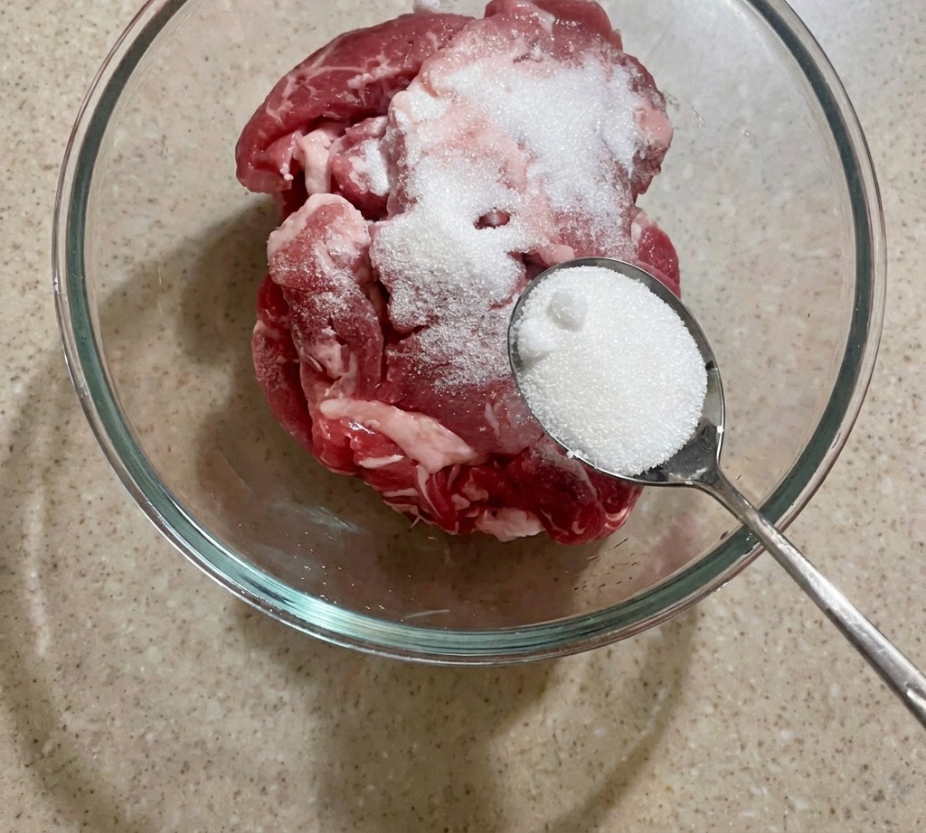 Una ciotola con fette di maiale crudo condite con un cucchiaio di zucchero bianco.
