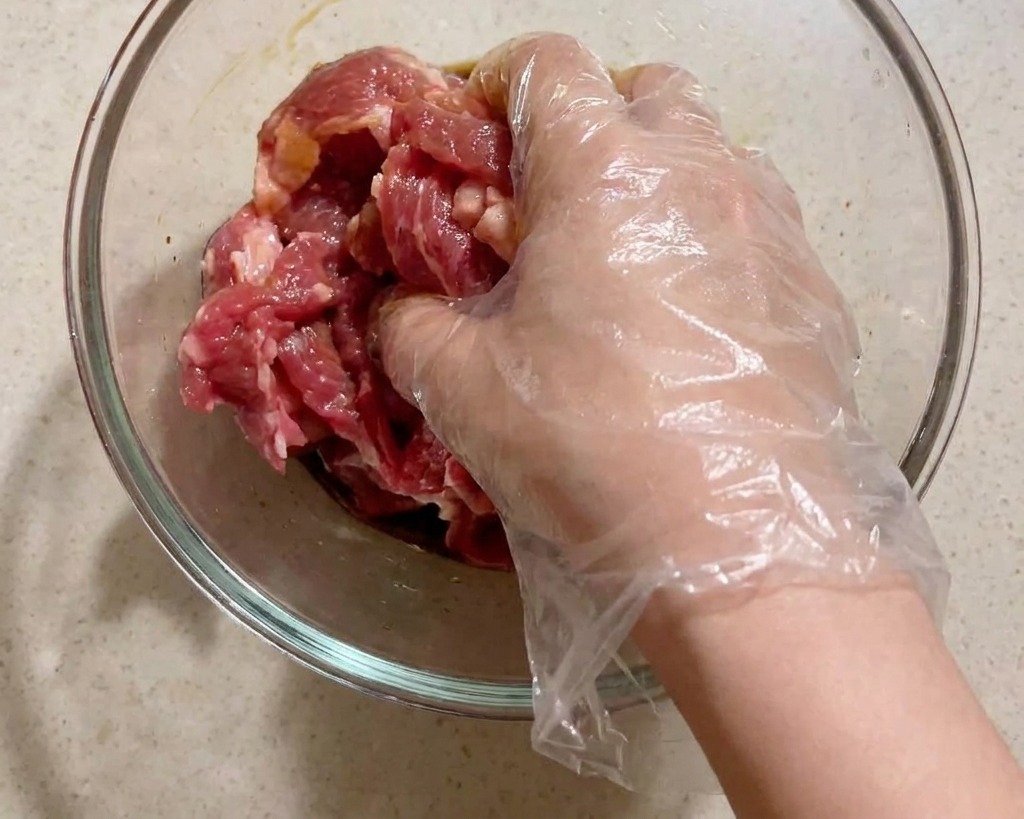 Una mano con un guanto in plastica che mescola fette di maiale crudo con condimenti liquidi in una ciotola di vetro.