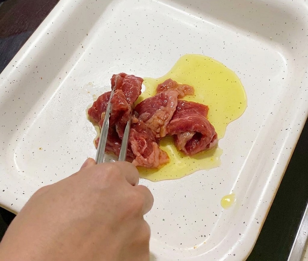 Pinze che adagiano fette di maiale marinato in una padella bianca con olio caldo.