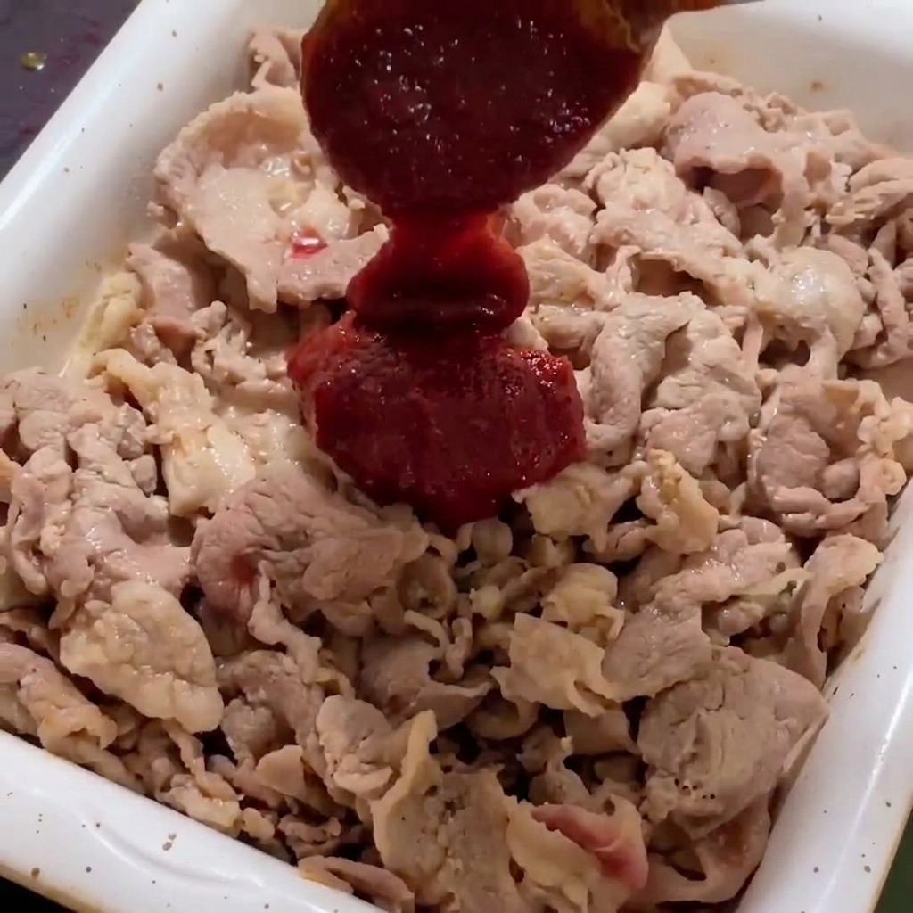 Salsa densa e rossa aggiunta alle fette di maiale dorate in una padella bianca.