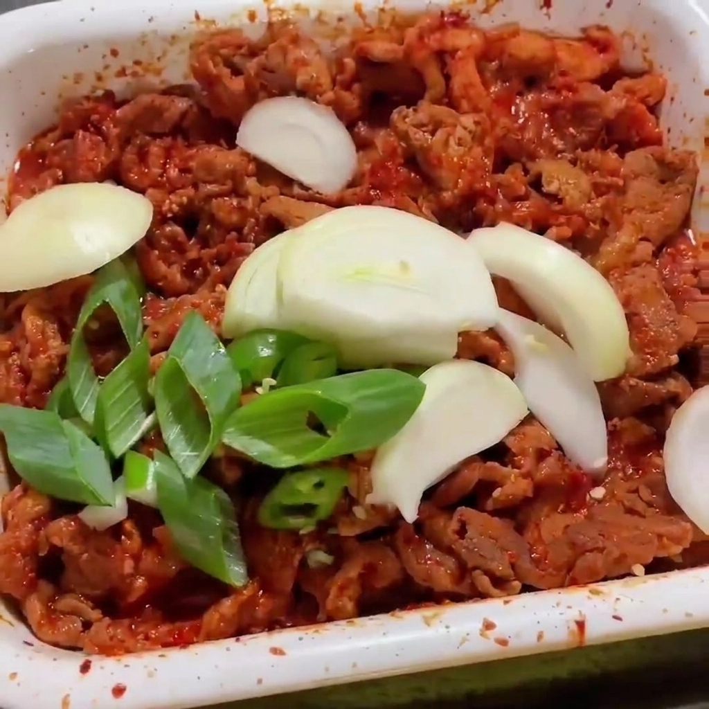 Cipolle bianche e cipollotti a fette sopra strisce di maiale piccante che sfrigolano in una padella bianca.