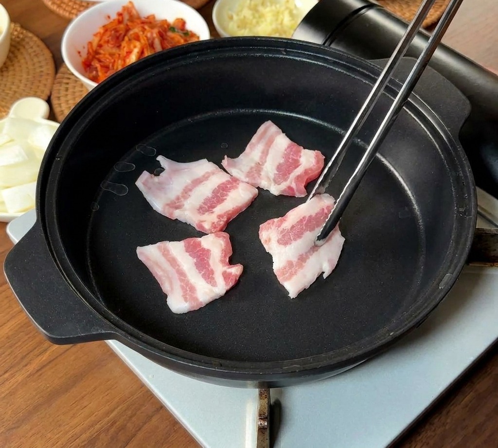Sottili strisce di pancetta di maiale marmorizzata inserite in una padella nera con lunghe bacchette di metallo.