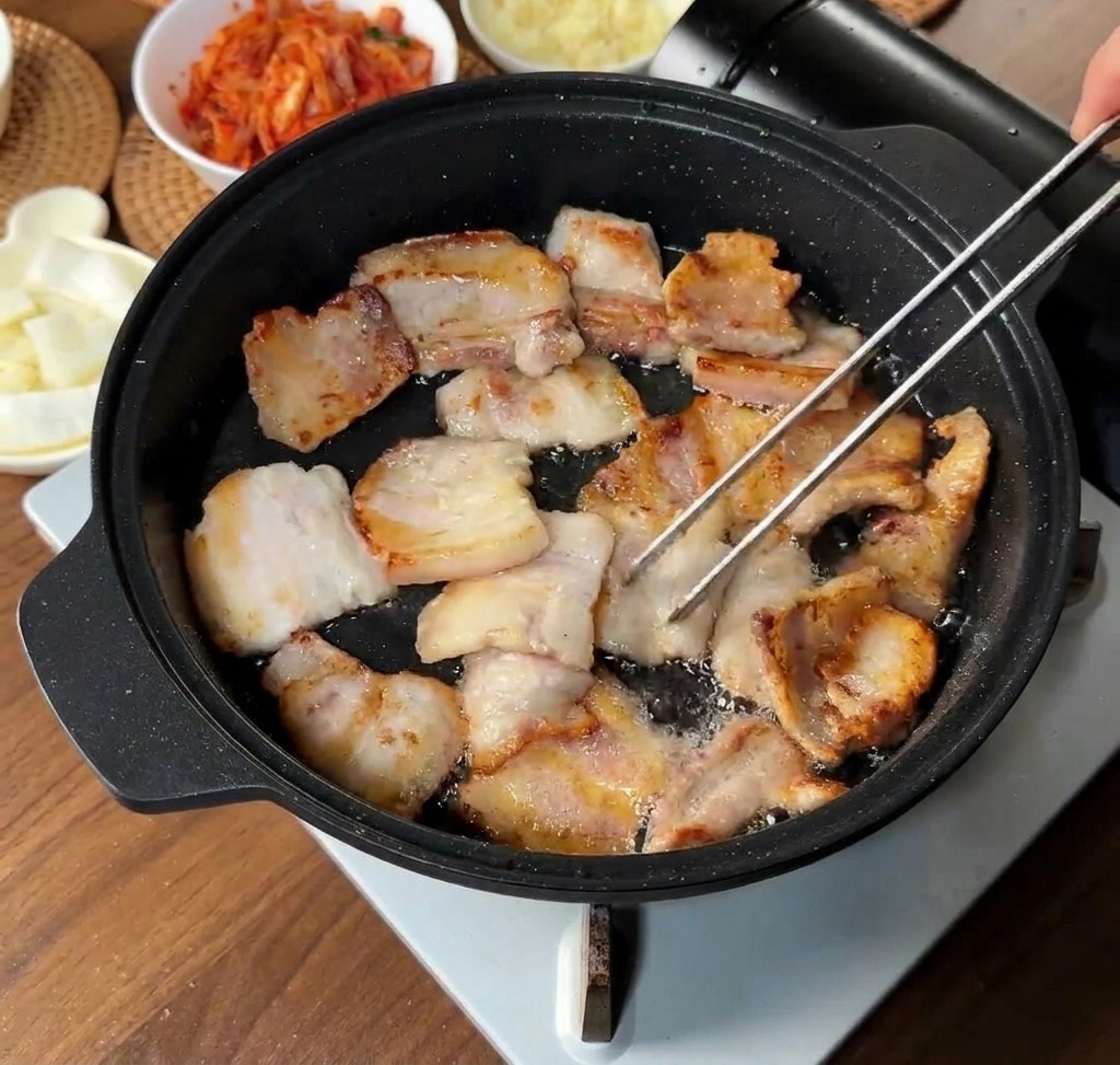 Fette di pancetta che rosolano nel loro grasso sciolto in una padella nera, con alcune fette girate dalle pinze.