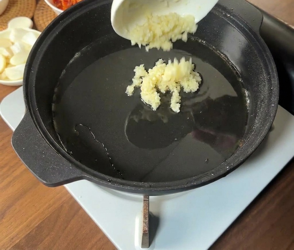 Aglio tritato versato da una piccola ciotola bianca nel centro caldo e oleoso di una padella nera.