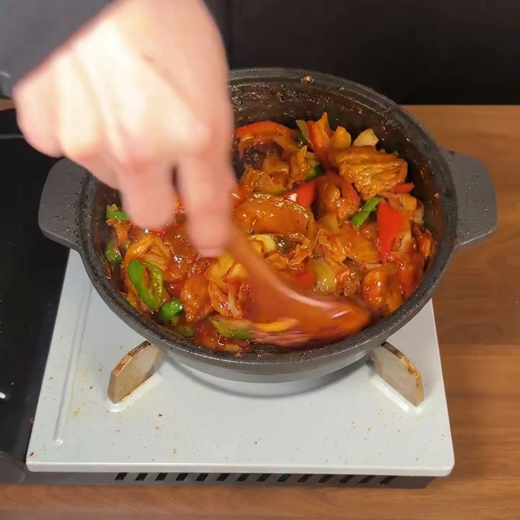 Un cucchiaio di legno che mescola energicamente una padella di pancetta coreana, kimchi e peperoncini in una densa salsa rossa.