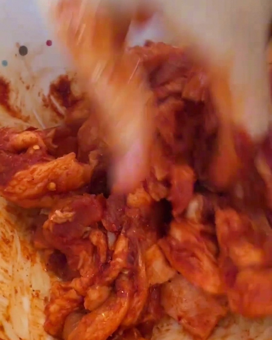 Una mano con guanto in plastica che mescola energicamente la carne marinata in rosso vivo in una ciotola bianca.