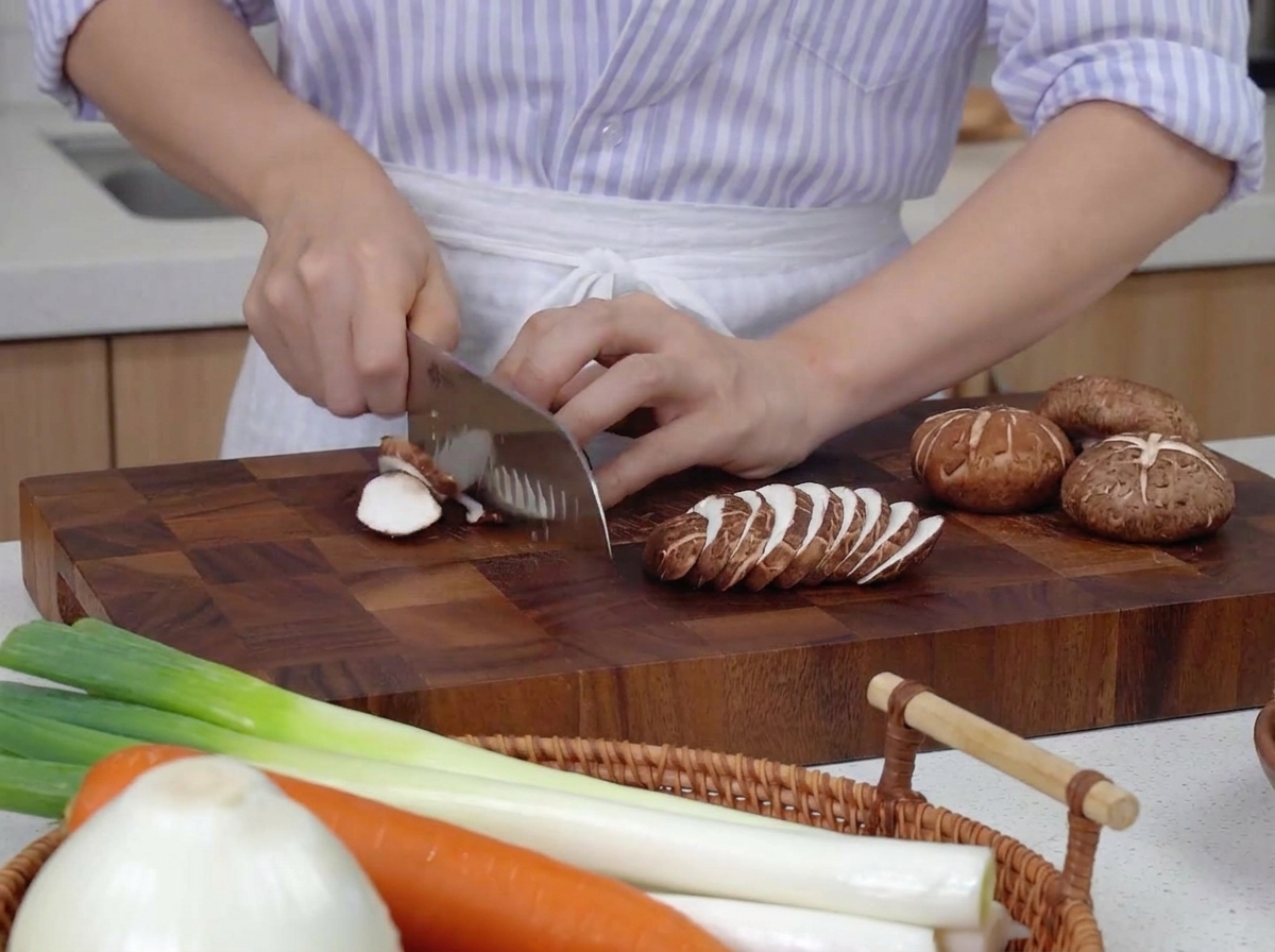 Mani che usano un coltello da chef per affettare sottilmente funghi shiitake marroni su un tagliere in legno scuro, accanto a carote intere e cipollotti.