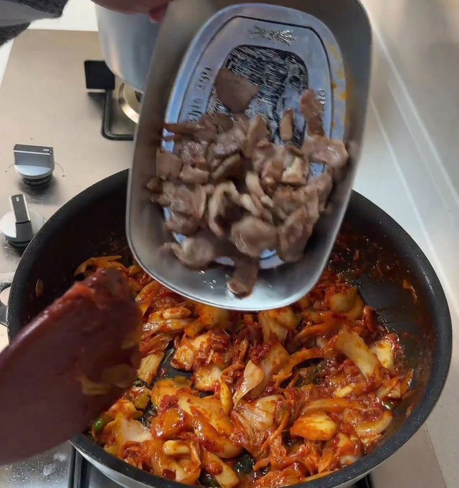 Fette di maiale scottate vengono versate da un vassoio di metallo in una padella con il mix piccante di kimchi e cipolle.