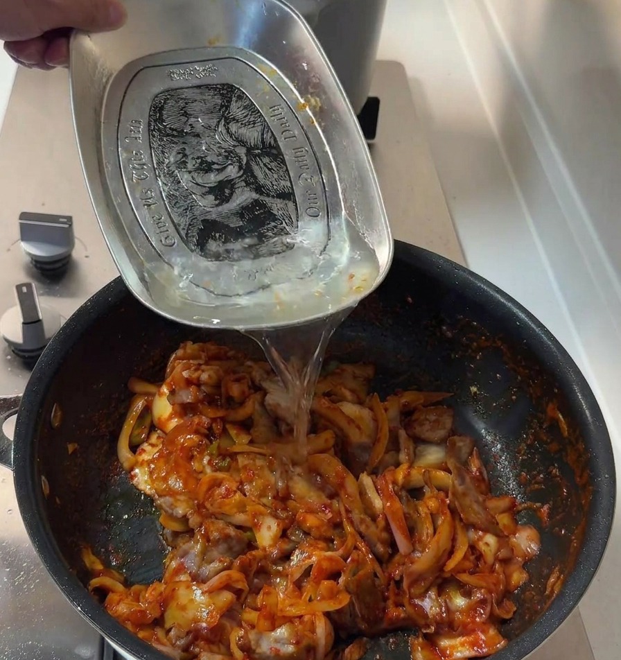 Lacqua viene versata da un vassoio di metallo nella padella con maiale e kimchi per completare la salsa.