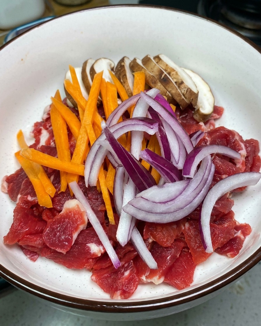 Una ciotola bianca con fettine di manzo crudo, funghi shiitake, carote a striscioline e cipolla rossa.