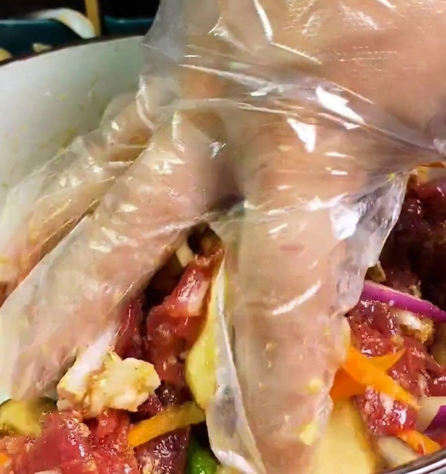 Una mano con guanto trasparente mescola manzo, cipolle, carote e funghi in una ciotola con marinata scura.