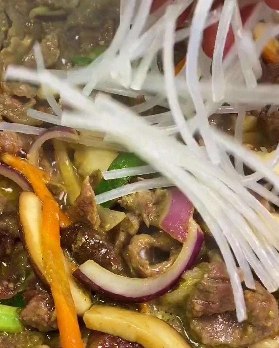 Una mano aggiunge una manciata di spaghetti di amido di patate dolci traslucidi in una padella di carne e verdure.