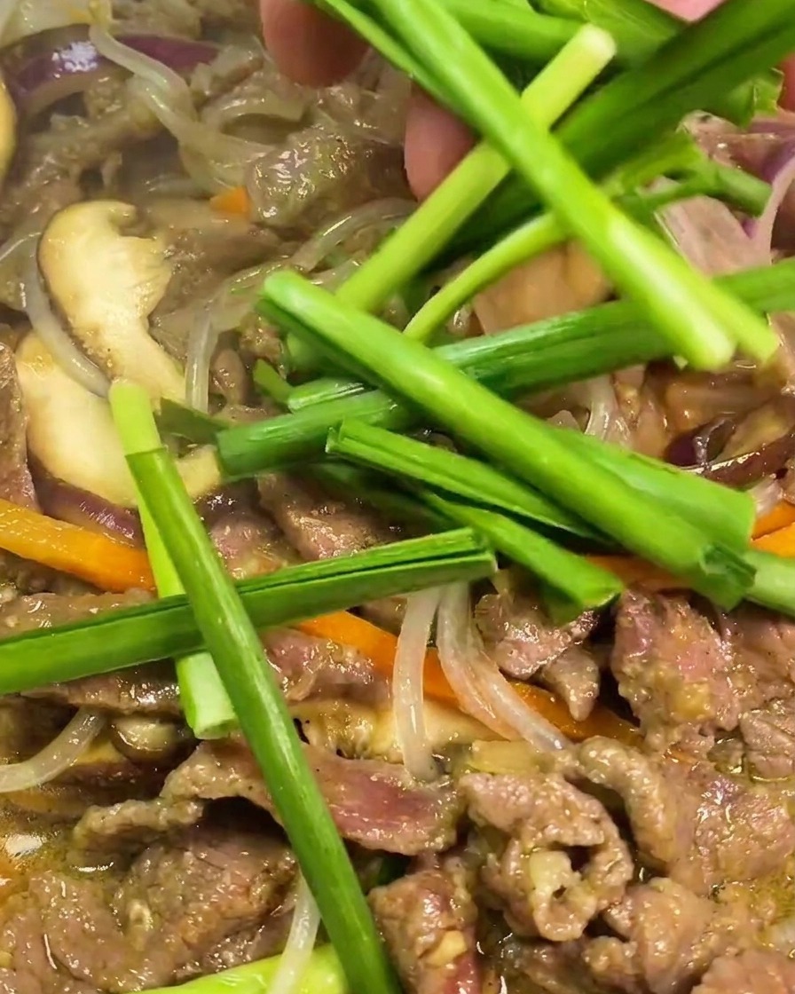 Segmenti di erba cipollina cinese aggiunti a mano in una padella calda con manzo, noodles traslucidi e verdure.