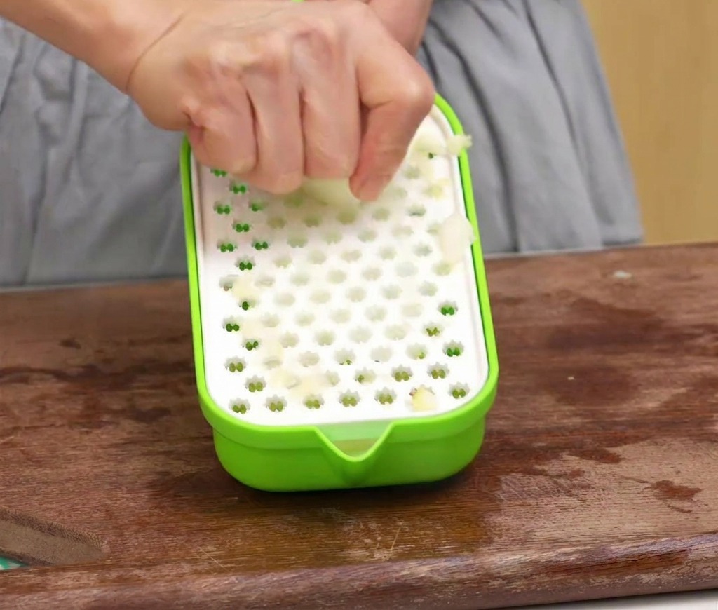 Una persona che usa una grattugia di plastica verde per trasformare una mela sbucciata in polpa su un tagliere di legno.