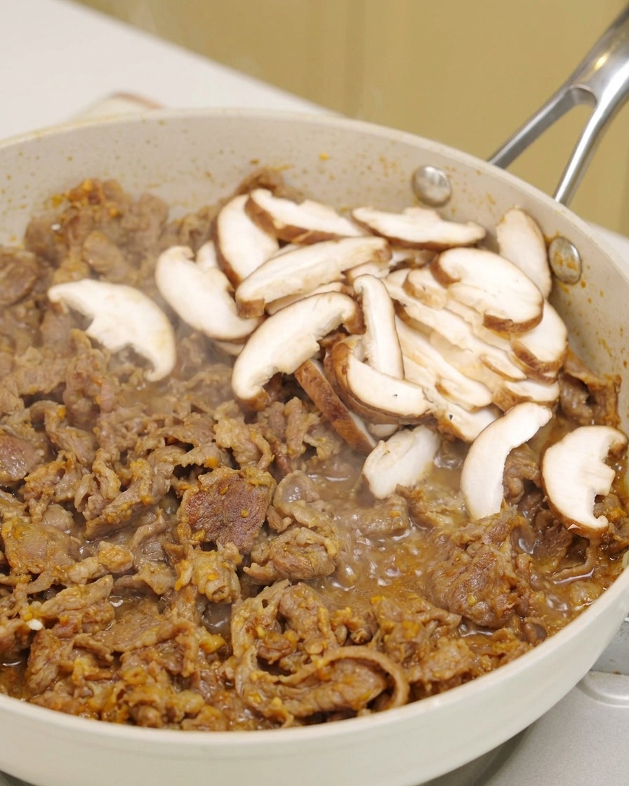 Fette di funghi shiitake freschi posizionate sopra il manzo sfrigolante in una padella bianca.