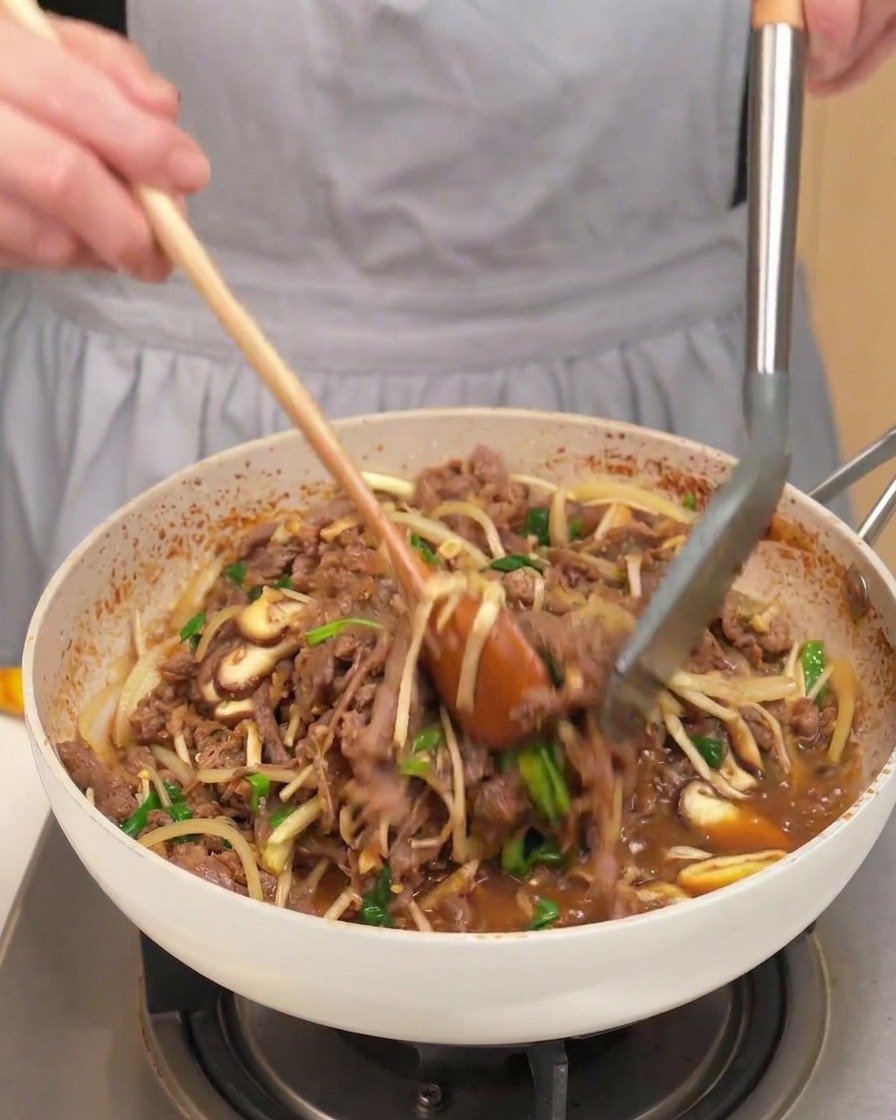Una padella piena di manzo cotto, funghi, cipolle e cipollotti saltati con un cucchiaio di legno e pinze.