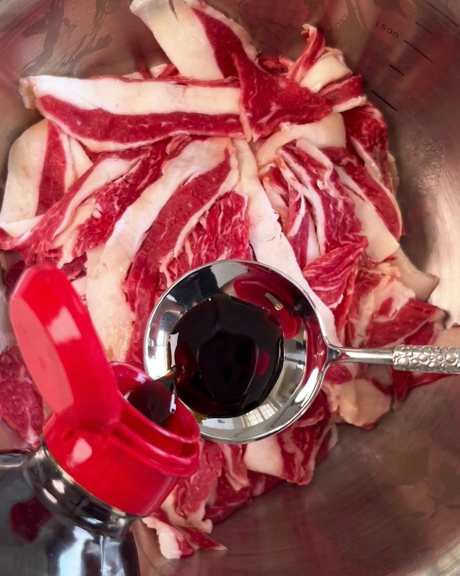 Salsa di soia scura versata da un cucchiaio di metallo su fette di manzo crudo in una ciotola dargento.