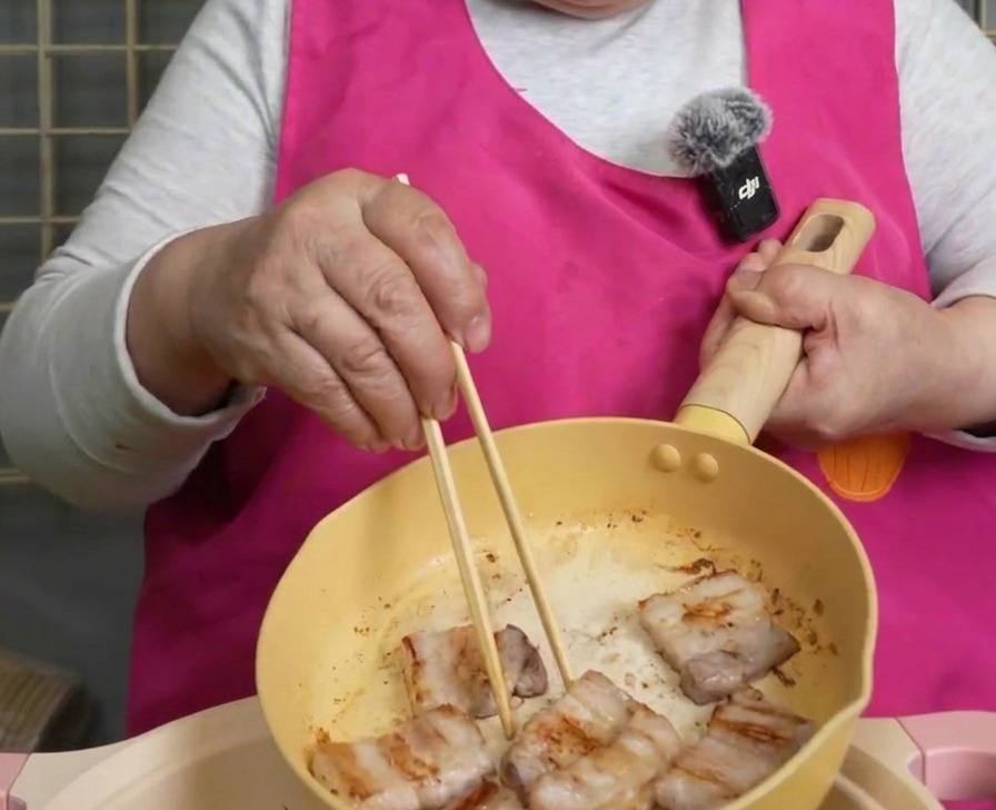 Fette di pancetta dorata sollevate dalla padella con bacchette di legno.