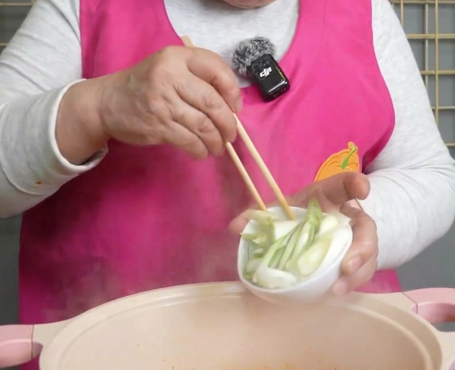 Cipollotti freschi tritati versati da una ciotolina in una pentola fumante di zuppa di kimchi.