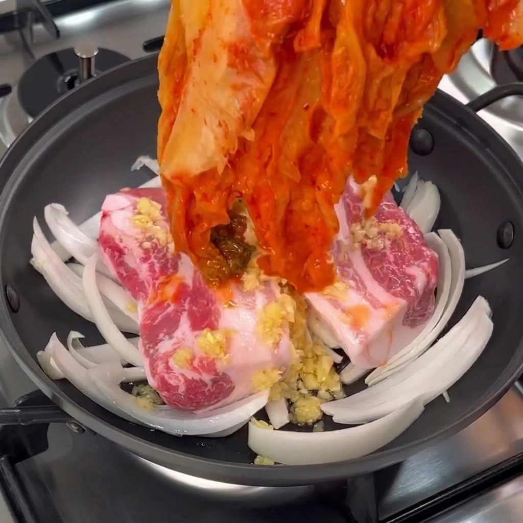 Una mano che adagia un grosso cespo di kimchi rosso invecchiato sopra i pezzi di pancia di maiale in padella.