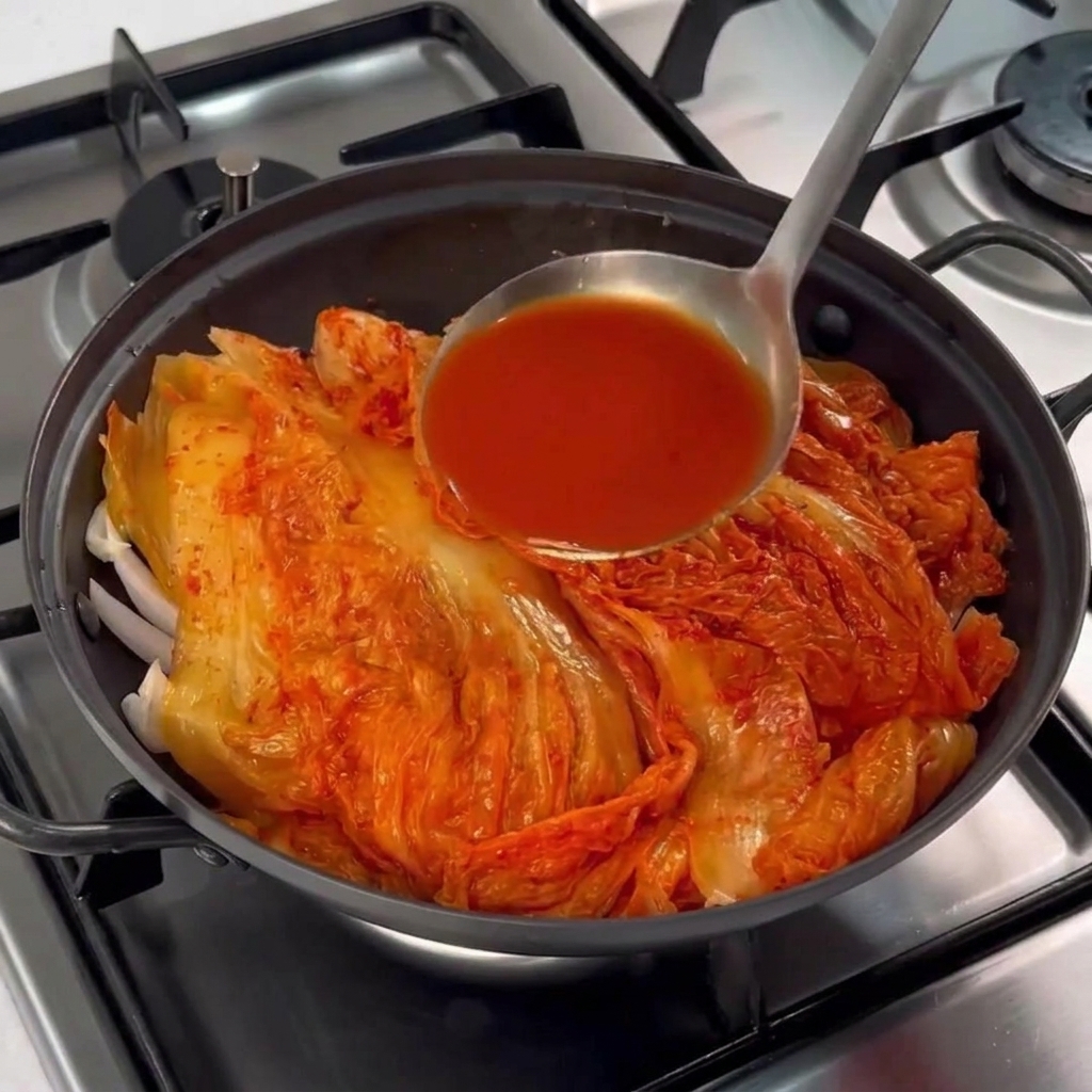 Un mestolo di metallo che versa la salamoia rossa del kimchi sopra il cumulo di verdure e carne.