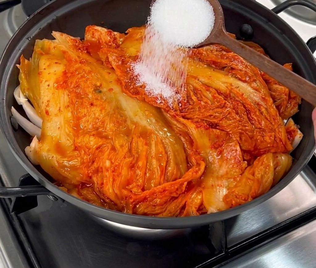Zucchero semolato bianco versato da un cucchiaio di legno sopra il kimchi rosso nella pentola.