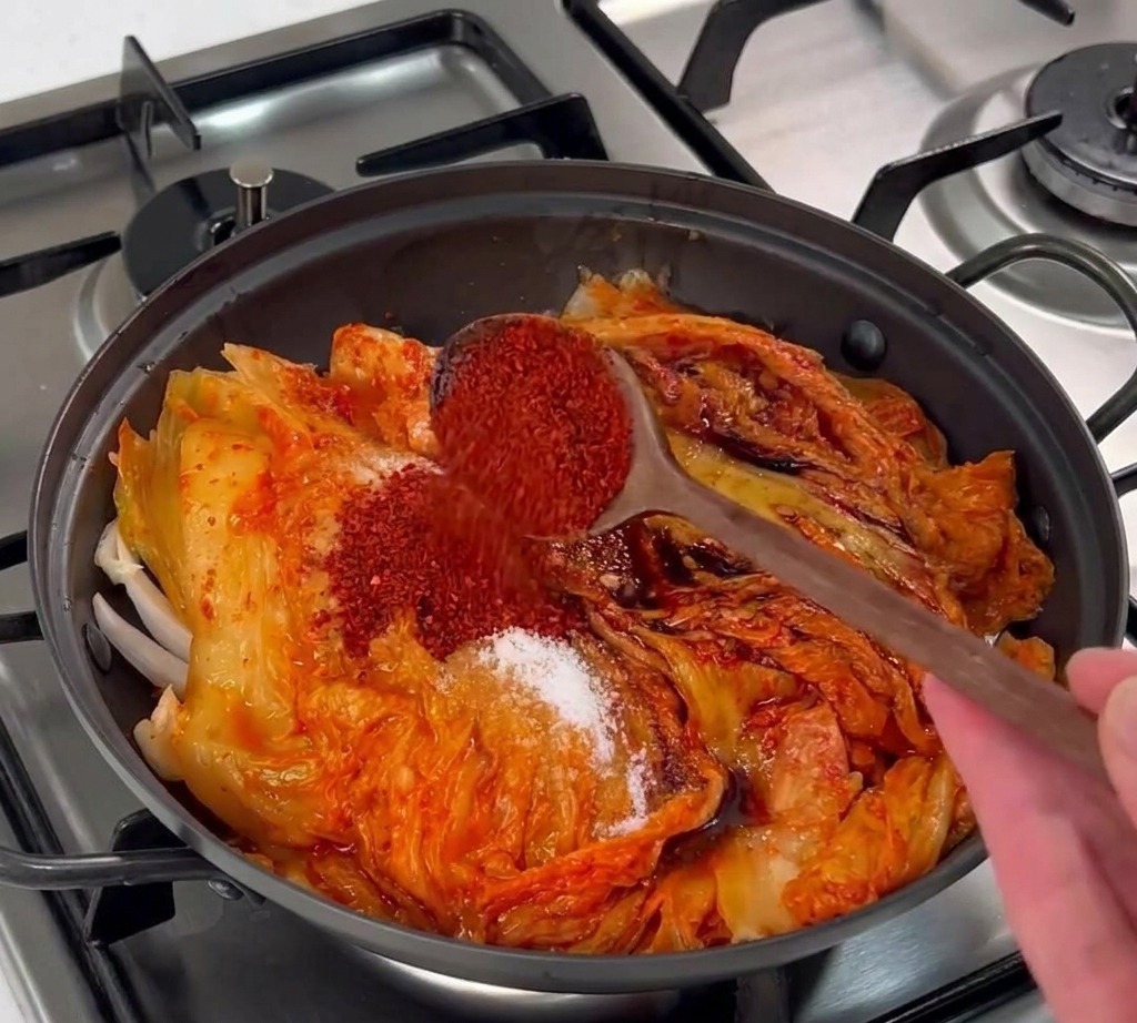 Un cucchiaio di legno che versa fiocchi di peperoncino coreano rosso acceso sopra il kimchi.