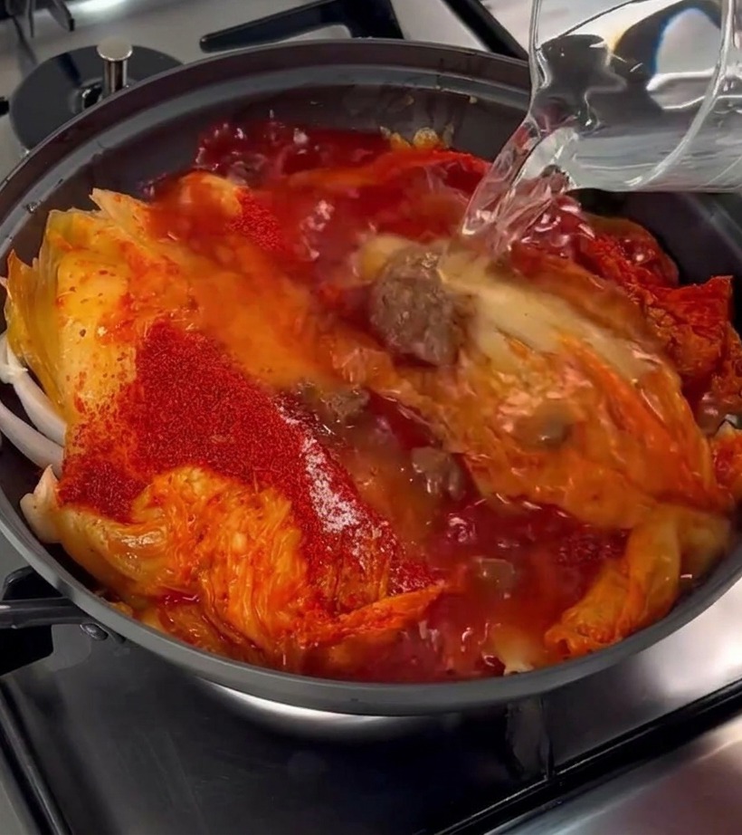 Acqua limpida versata da una caraffa di vetro in una pentola contenente kimchi, maiale e peperoncino.