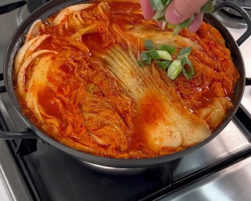 Una mano che sparge cipollotti freschi tritati sopra una pentola colma di kimchi e brodo rosso.