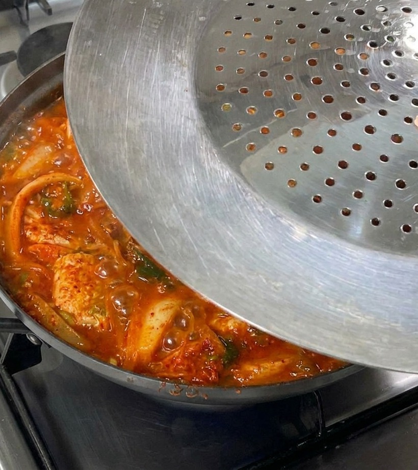 Una mano che posiziona un coperchio di metallo sopra la padella fumante di stufato su un fornello a gas.