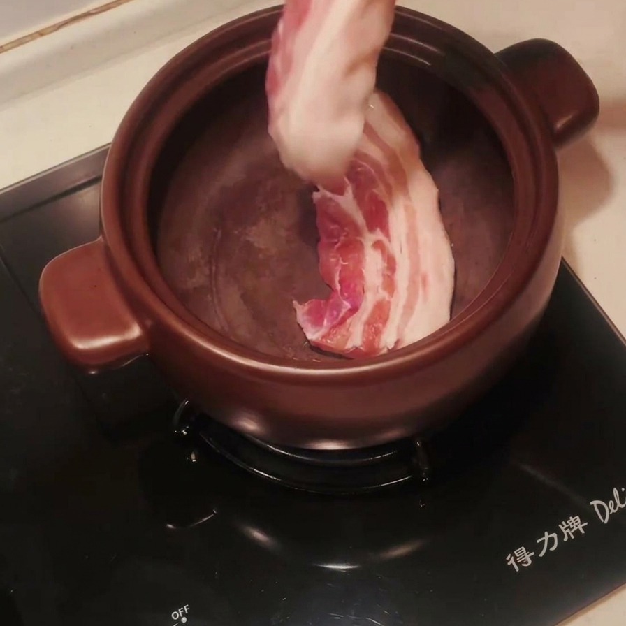 Pancetta di maiale cruda a strisce messa in una pentola di terracotta scura preriscaldata con delle pinze.