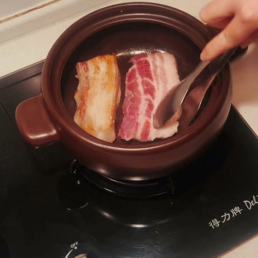 Strisce di pancetta nella pentola di terracotta girate con le pinze, con un lato perfettamente dorato.