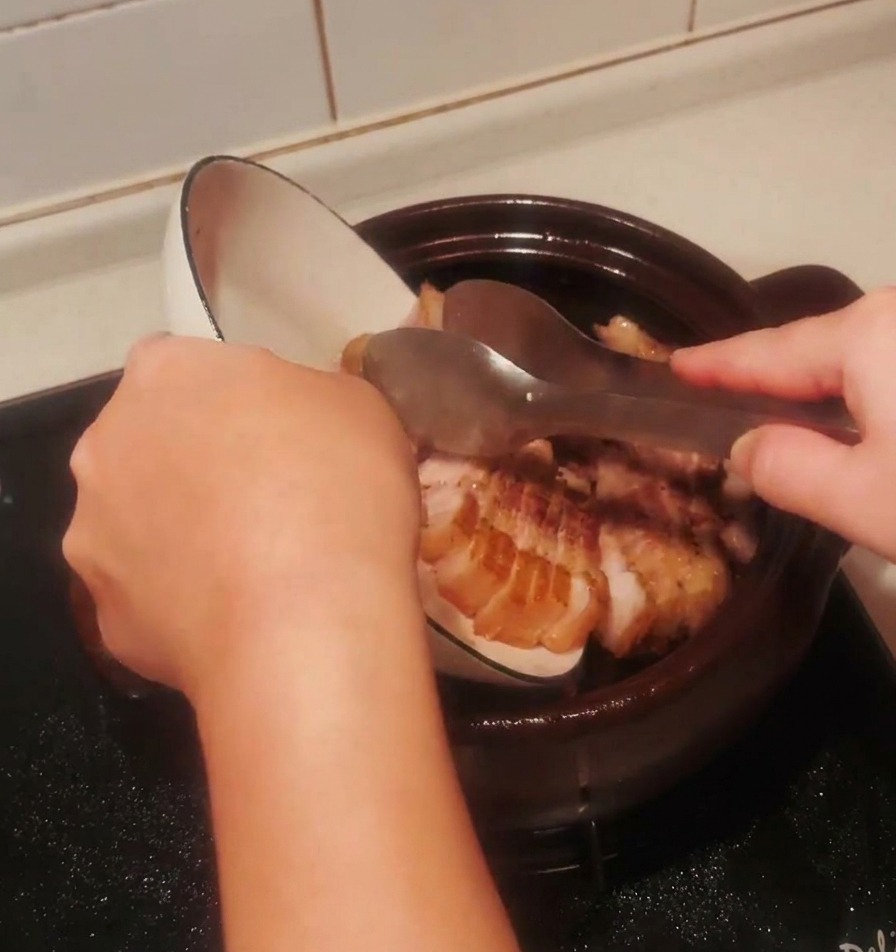 Pezzi di pancetta rosolati versati da una ciotola bianca di nuovo nella pentola di terracotta scura.