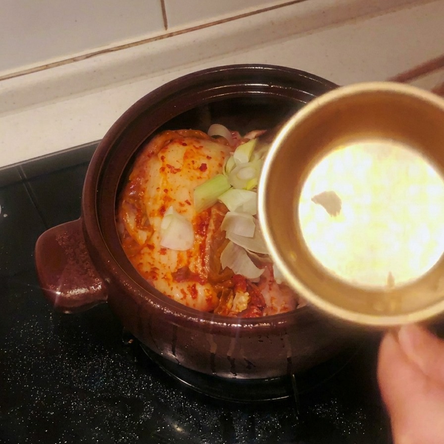 Una ciotola di metallo dorata che versa aglio affettato e grossi segmenti di cipollotto sul kimchi nella pentola.