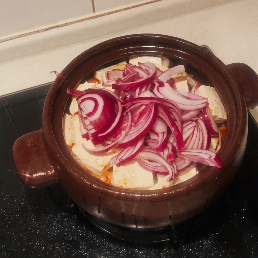 Un mucchio di cipolle rosse affettate che copre lo strato di tofu bianco nella pentola di terracotta.