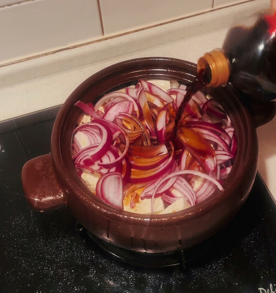 Salsa di soia scura versata da una bottiglia su cipolle rosse e blocchi di tofu nella pentola.