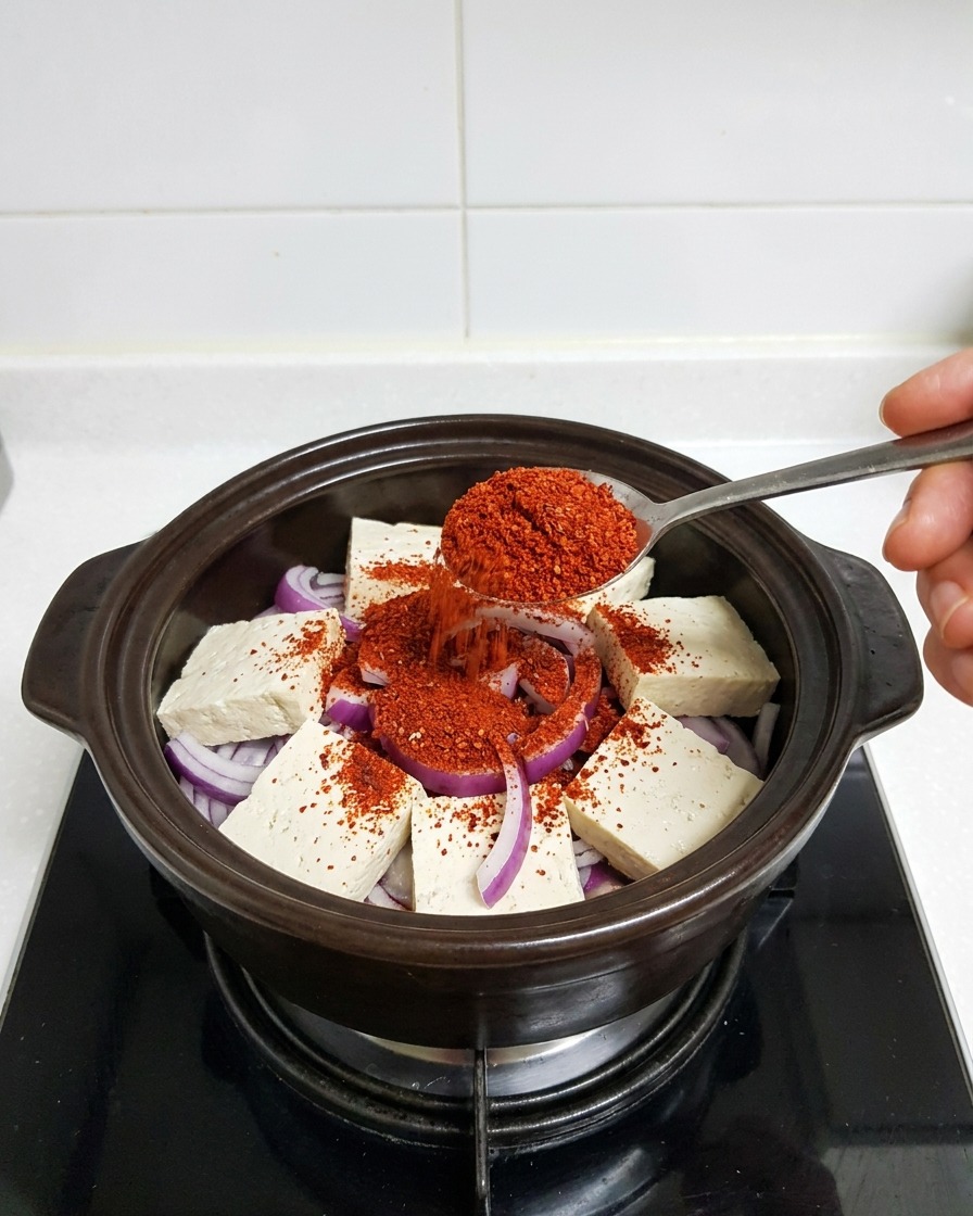 Un cucchiaio di metallo che mette peperoncino rosso fine su tofu e cipolle nella pentola di terracotta.