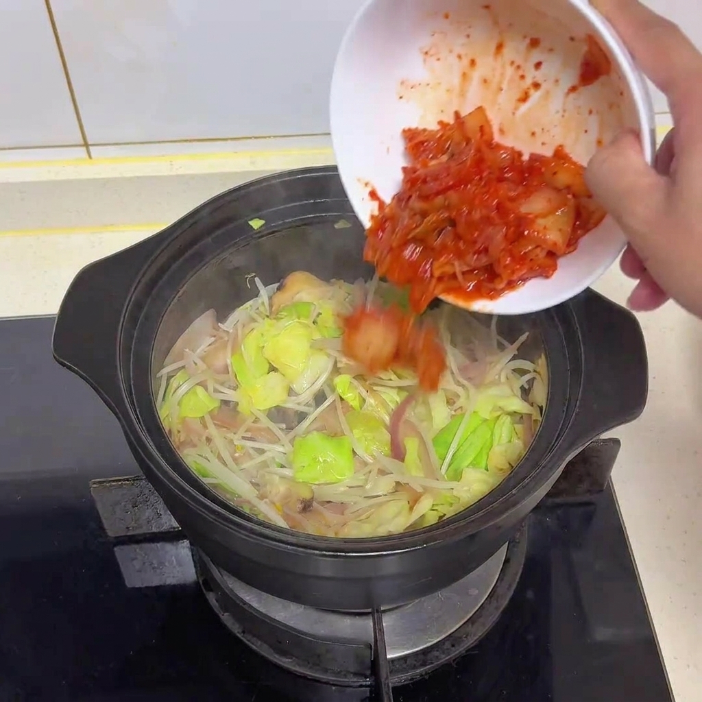 Kimchi rosso acceso tritato versato da una ciotola bianca in una pentola con verdure soffritte.