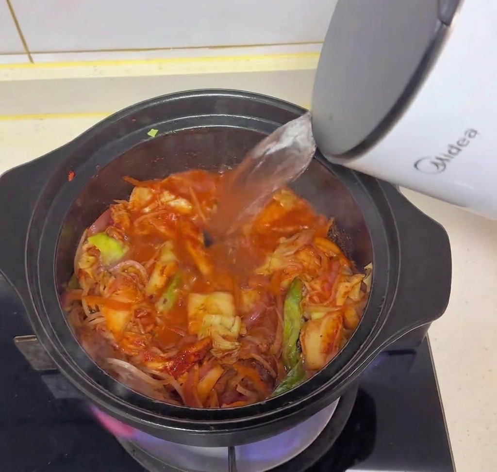 Acqua calda versata da un bollitore elettrico bianco nella pentola nera con kimchi e verdure soffritte.