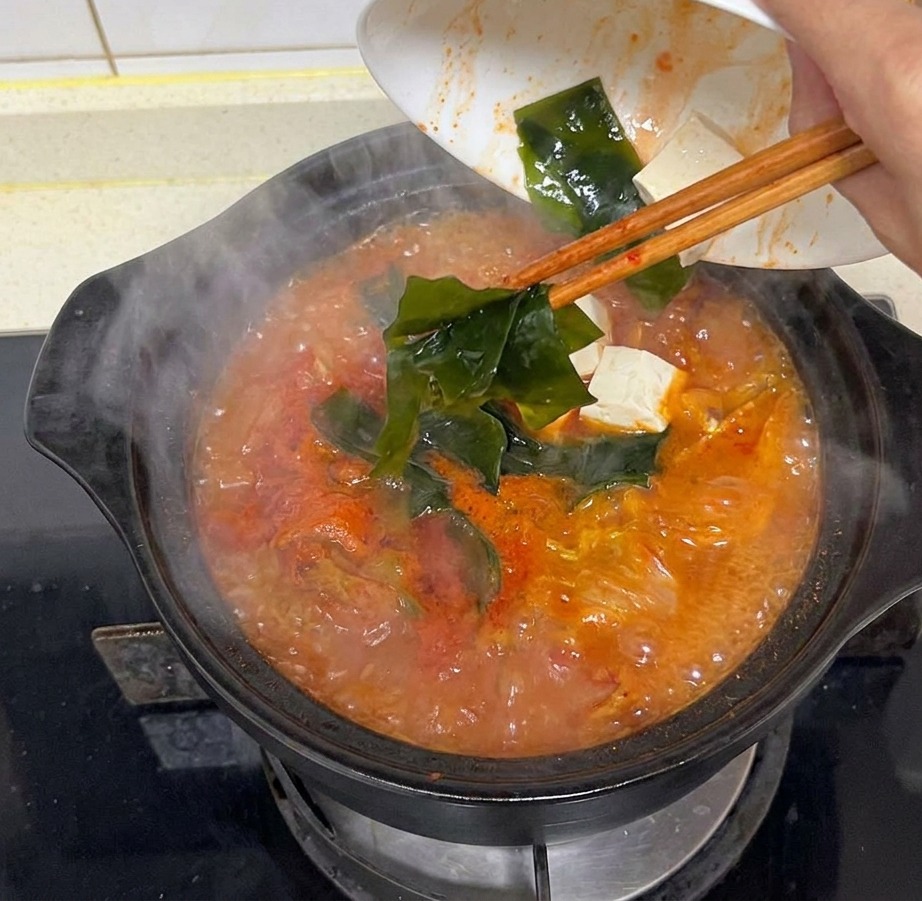Una mano versa pezzi di alga verde scura in una pentola fumante di zuppa di kimchi rossa sul fuoco.