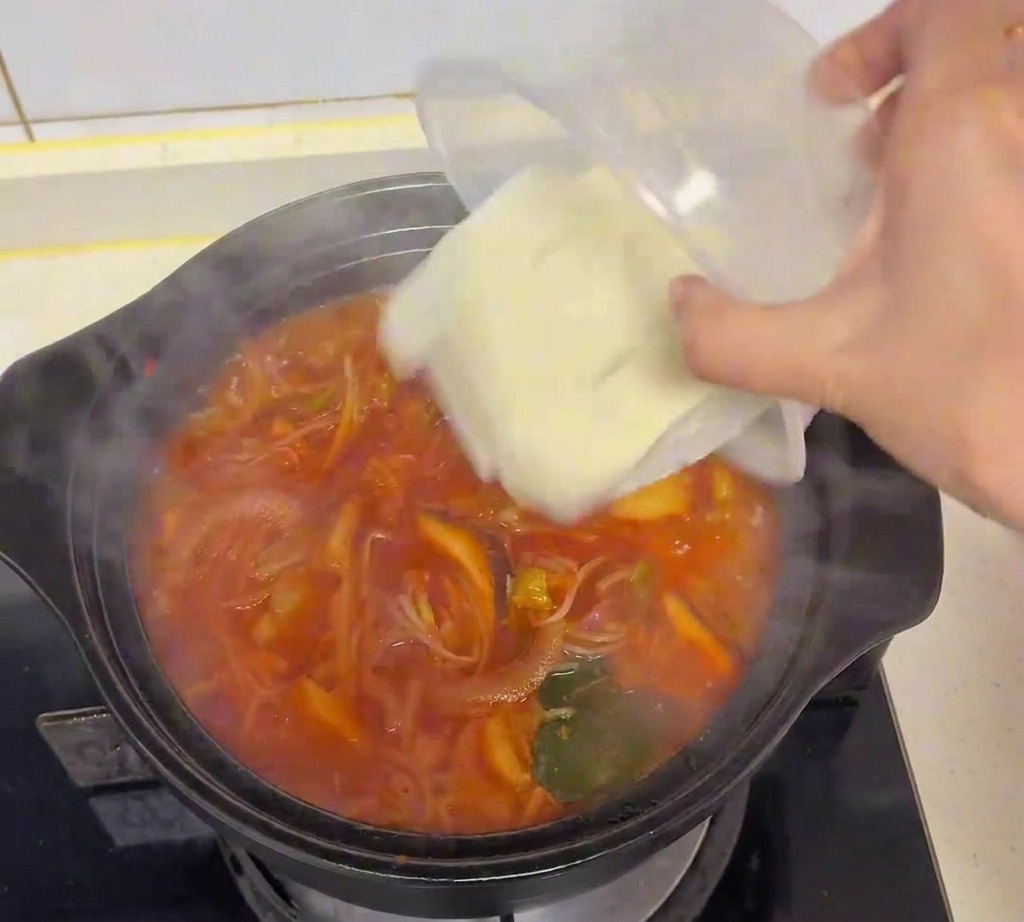 Cubetti di tofu bianco versati da un contenitore di plastica trasparente nella zuppa di kimchi bollente.