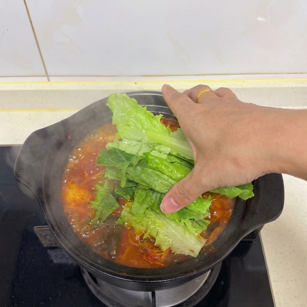 Una mano sistema foglie di lattuga verde sopra la zuppa di kimchi e tofu fumante.
