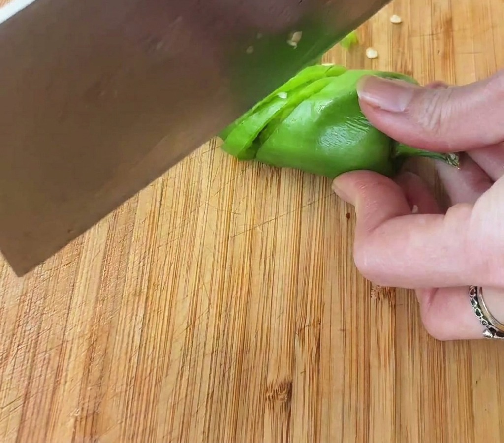 Primo piano di una mano che usa una mannaia per affettare un peperoncino verde brillante a rondelle sottili.