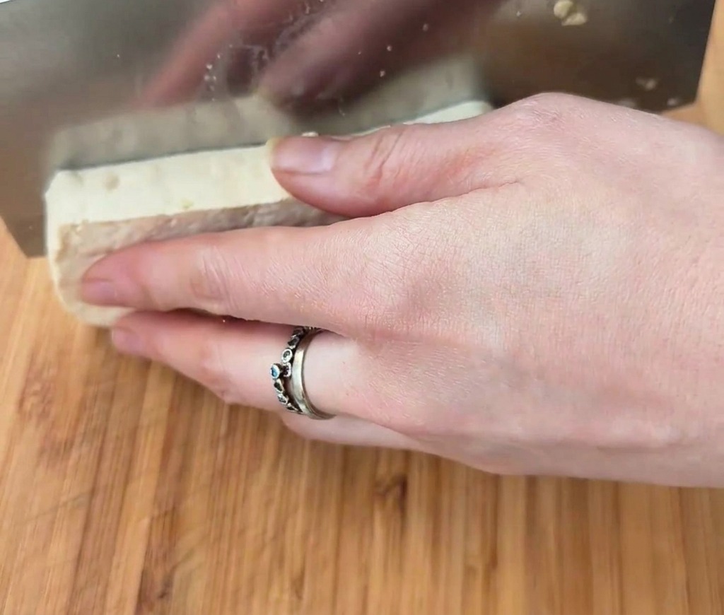 Un blocco rettangolare di tofu bianco tagliato a cubetti con una mannaia di metallo su una superficie di legno.