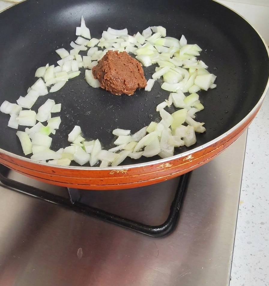 Una cucchiaiata di densa pasta di soia marrone scuro aggiunta a una padella con cipolle parzialmente soffritte.