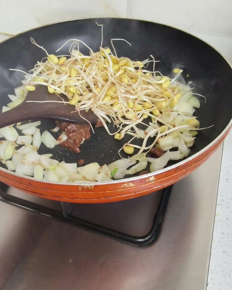 Una spatola di legno che mescola germogli di soia, cipolle e pasta di soia in una padella arancione.