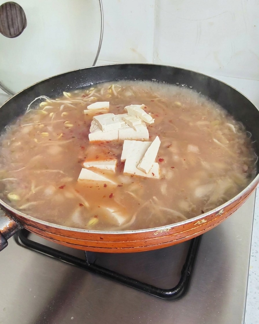 Cubetti di tofu che riposano sulla superficie di una zuppa rossastra che sobbolle in una grande padella arancione.