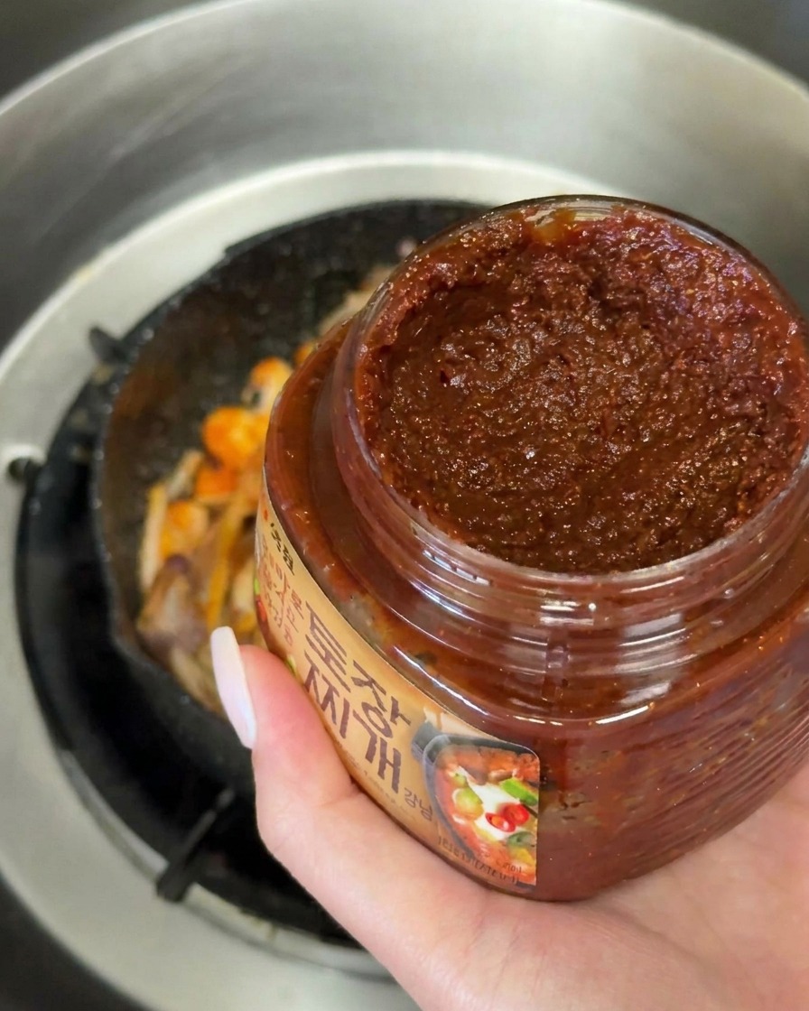 Una mano aggiunge un cucchiaio colmo di pasta di soia coreana scura alle verdure in padella.